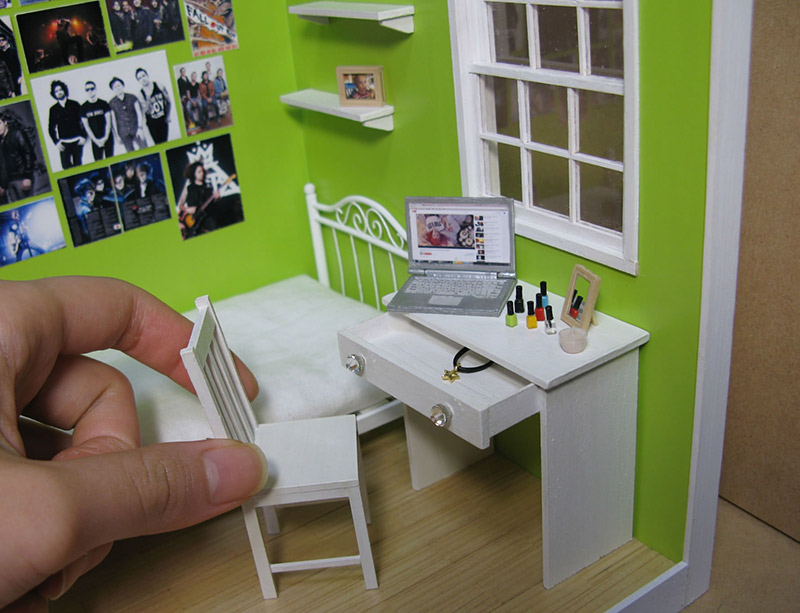 A normal-sized hand pulling out a chair from a small desk in a miniature room