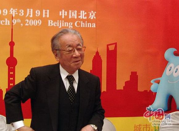 A Japanese man in a suit with glasses against a stylized Beijing skyline background