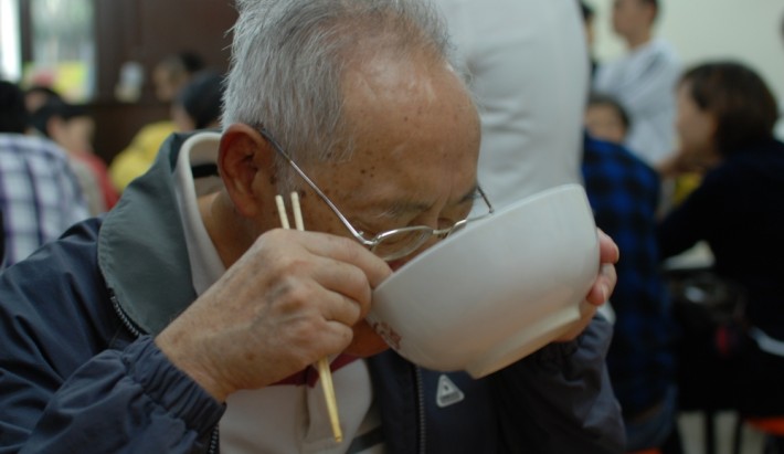 An elderly Japanese man lifting up his bowl of noodles