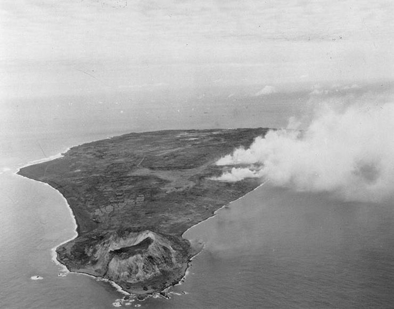 Black and white aerial image of a small island with a crater at one end and smoke rising from somewhere behind it