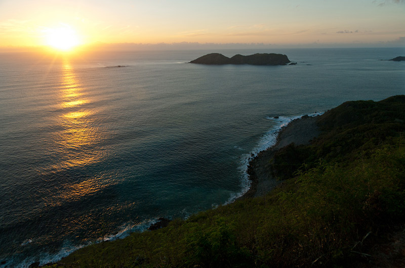 The sun rising over a calm sea with a bit of shoreline in frame