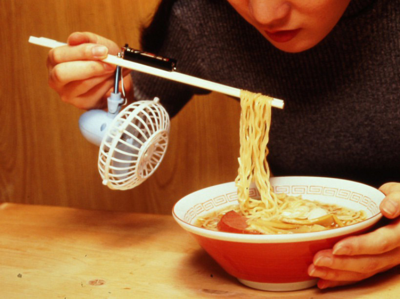 A person eating a bowl of ramen with a fan connected to a battery between the chopsticks blowing the ramen